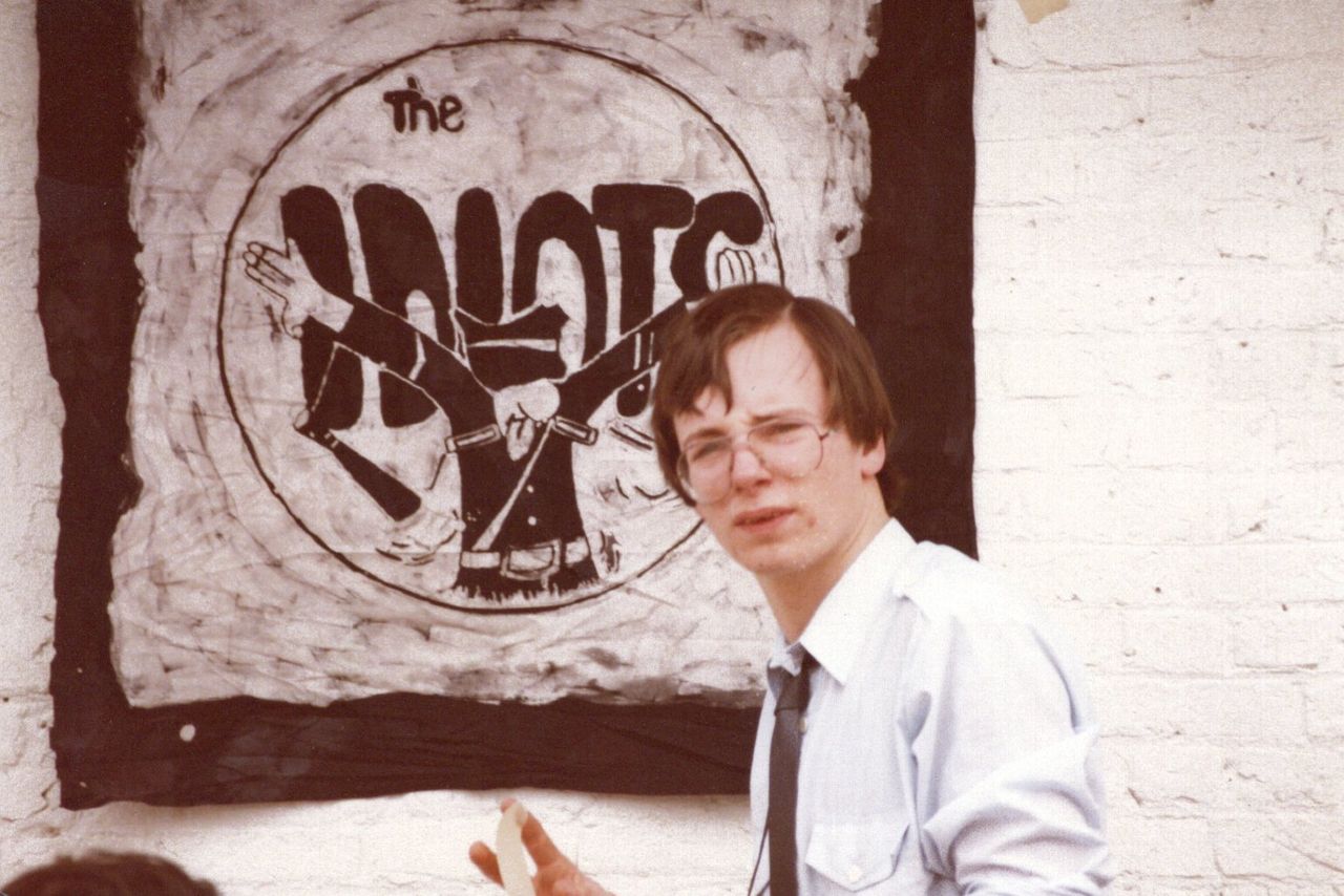 Hand-painted The Idiots banner on a whitewashed brick wall, with a bystander in front