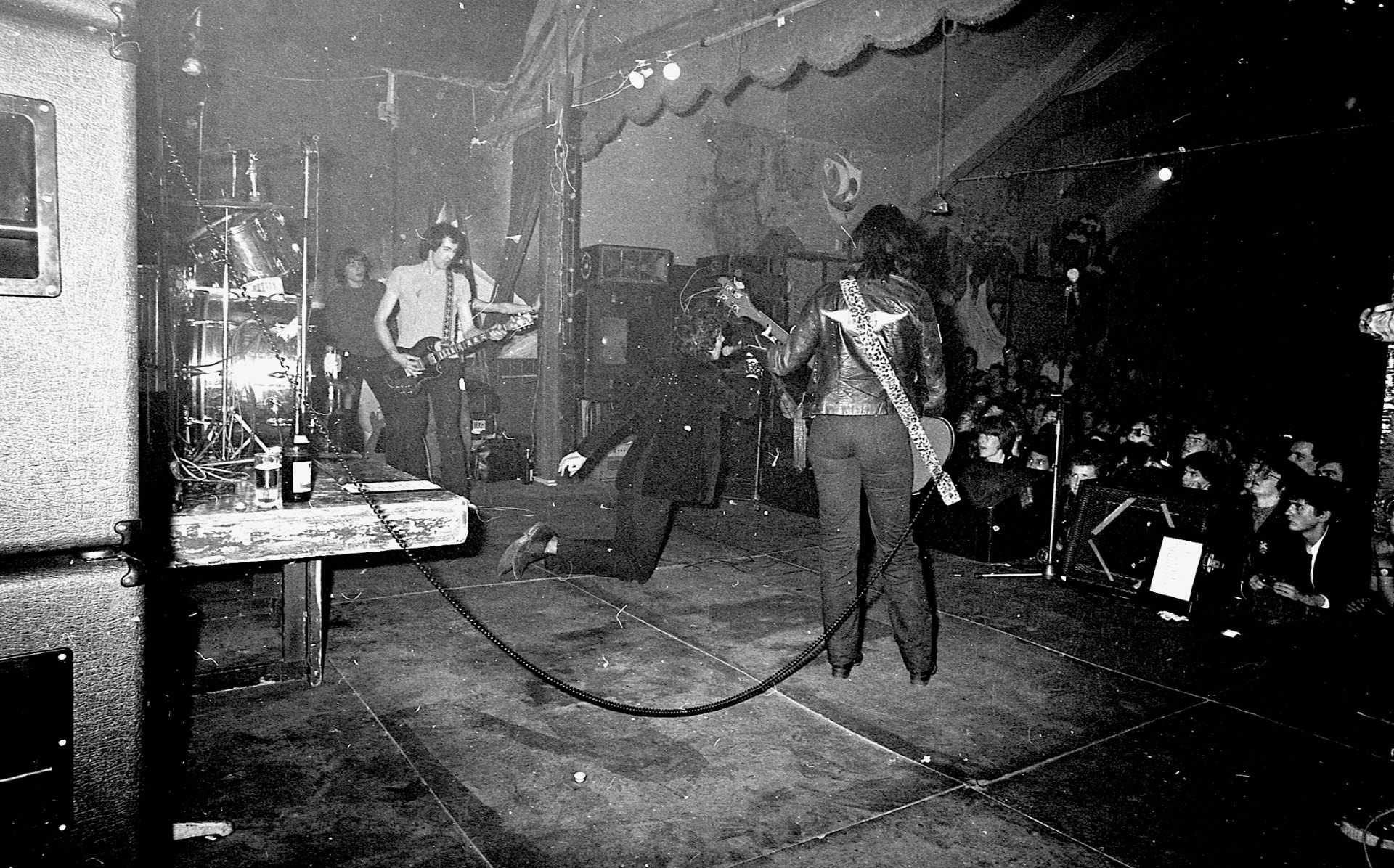 Black and white photograph of a band mid-set on a small club stage, singer on his knees, crowd at the side